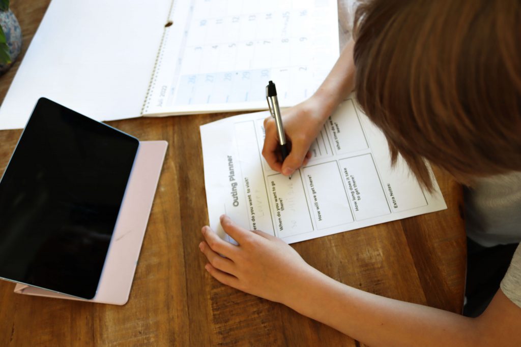Child filling out the Montessori outing planner worksheet. Using a tablet to research. In Montessori schools, elementary aged children are given the opportunity to organise, plan, and prepare for a trip outside of the classroom!